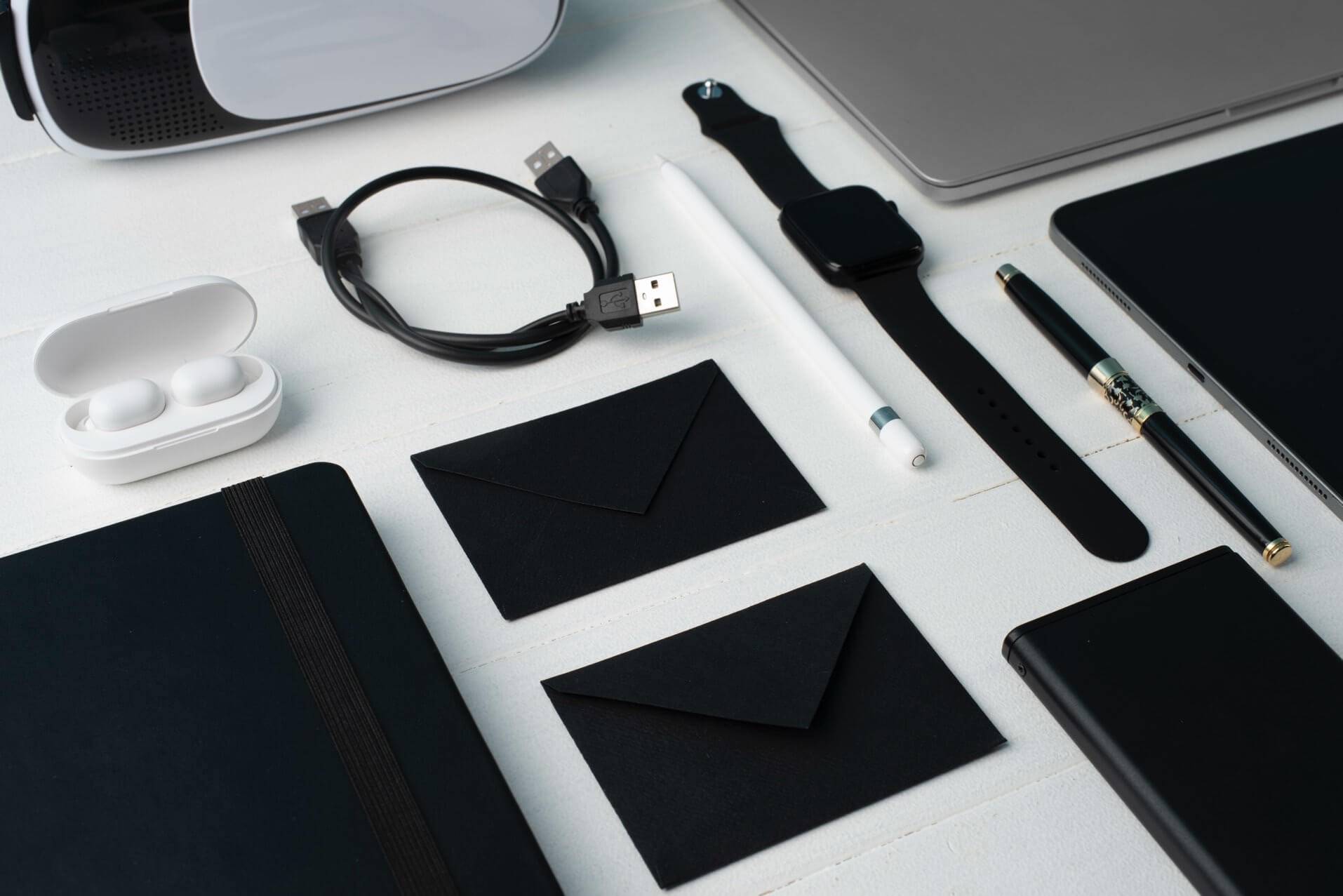 Modern stationery collection flat lay – black notebooks, wireless earbuds, smartwatch, USB cable and pens on white background